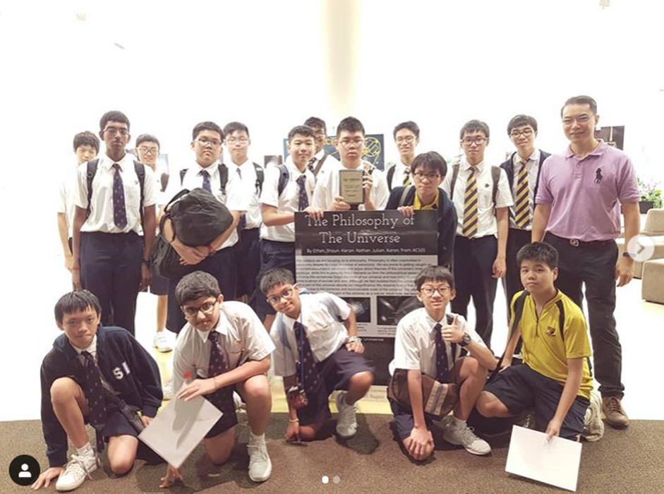 A large group of male students in white school uniforms and ties pose for a group photo, with one student holding a banner.